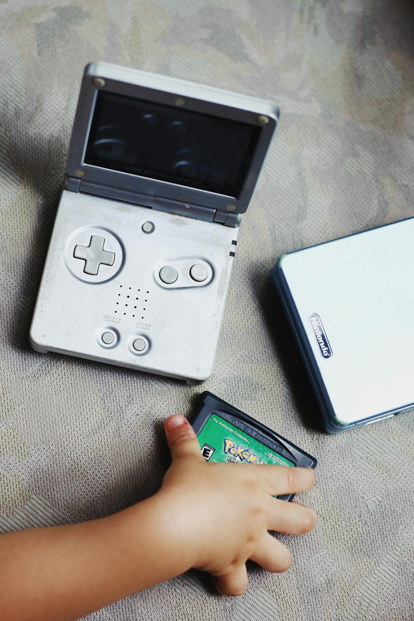 A child's hand holds a vintage Gameboy cartridge next to an open Gameboy SP console.